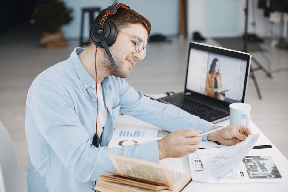 Hanley English - Online English Classes Man working remotely with headphones, reviewing documents, and video conferencing on laptop.